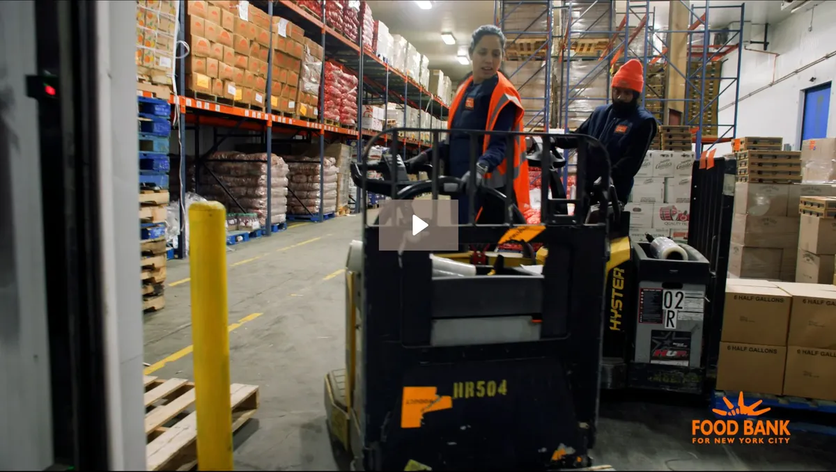 Warehouse workers operate a forklift amidst stacked food supplies and pallets in a busy food bank environment