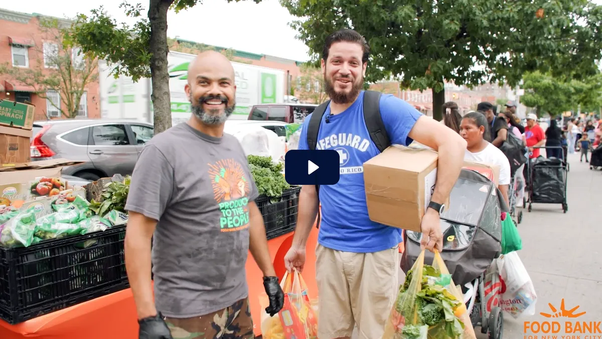 Two smiling men at an outdoor food distribution event, one wearing a 'Produce to the People' shirt and the other carrying a box and bags of fresh produce, with a line of people and food supplies in the background. Organized by Food Bank For NYC.