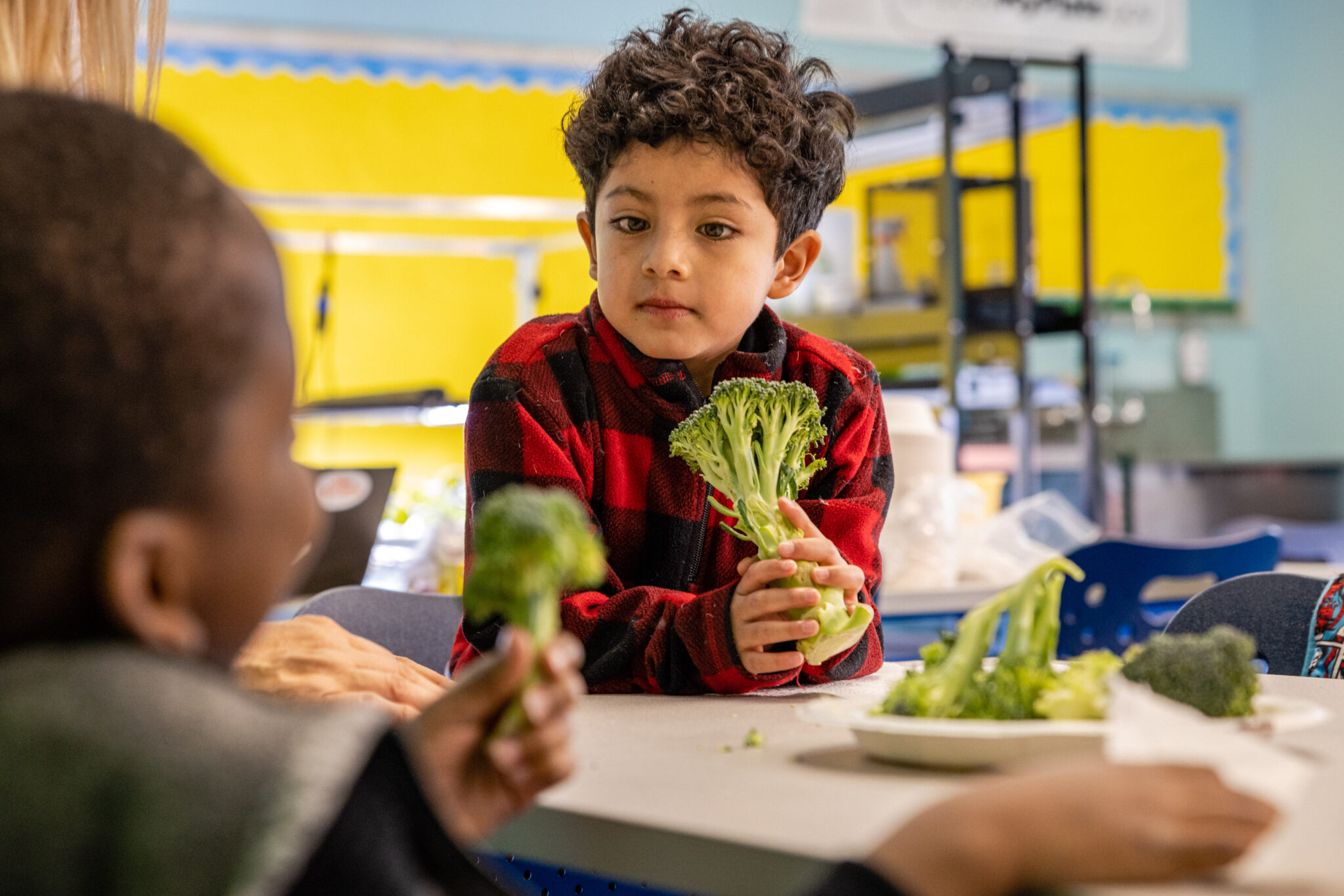 CookShop kid looks at another student while holding a large broccoli stem.
