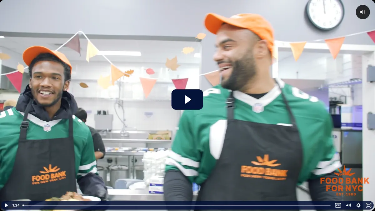Two volunteers in green jerseys and aprons serve food in a bustling kitchen decorated with colorful fall-themed banners.