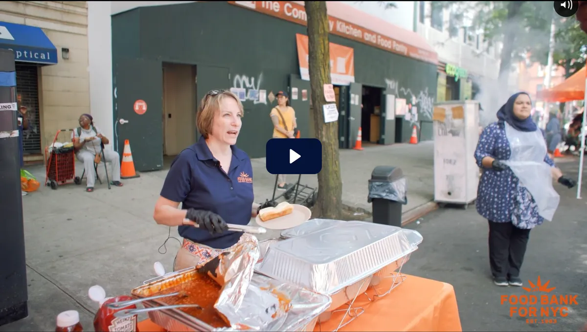 A volunteer (Leslie Gordon) serves food at a community event, with various people and food stations visible in an outdoor setting.