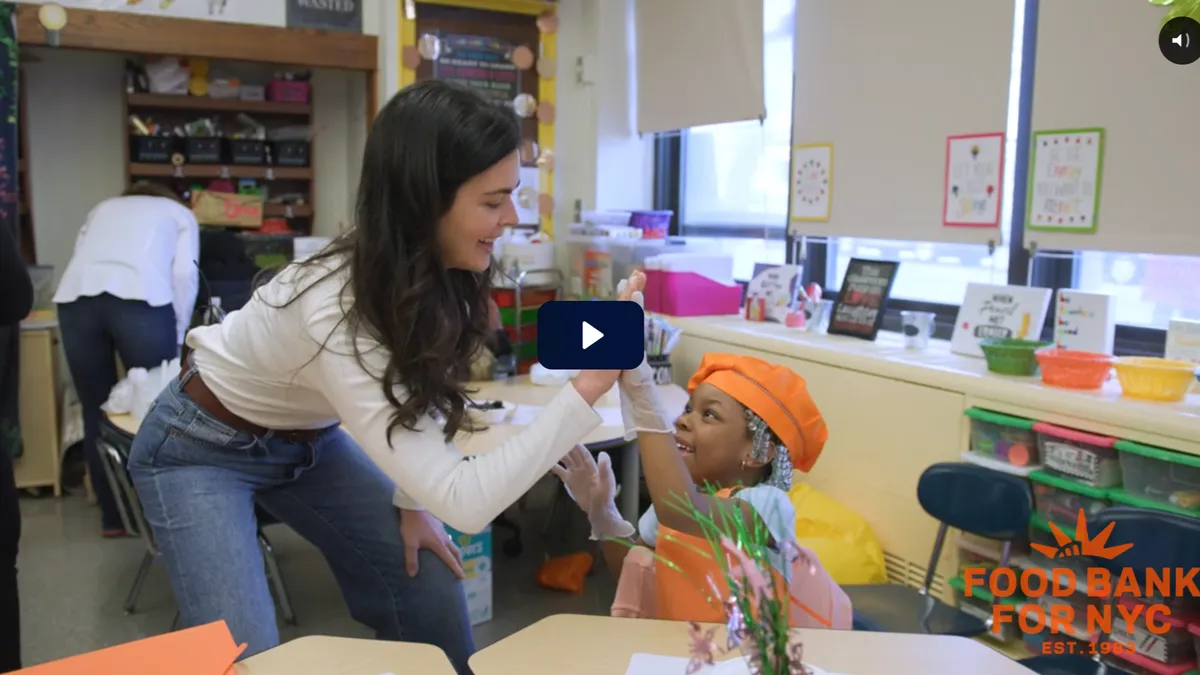 Woman giving a high five to a smiling young girl wearing an orange chef hat and gloves in a colorful classroom, during a Food Bank For NYC educational activity.