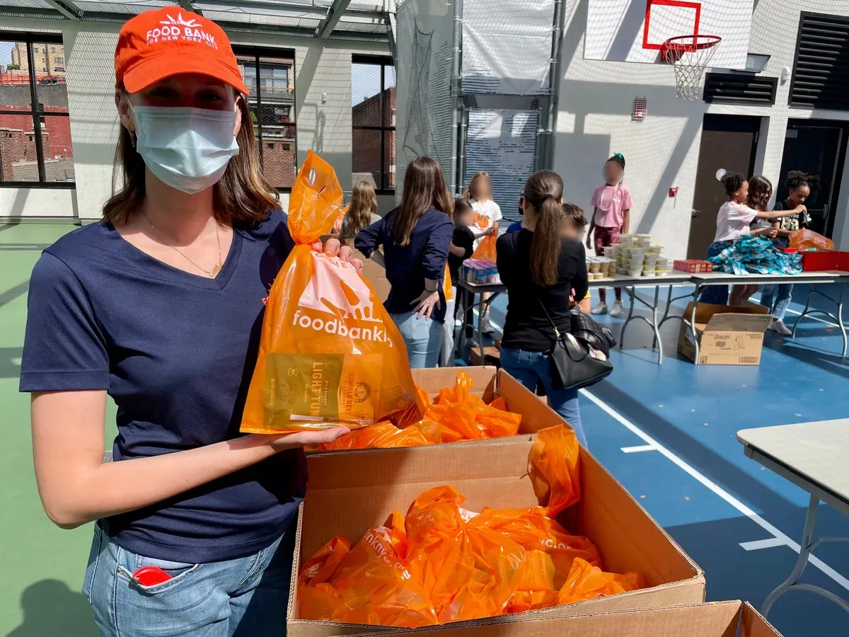 A volunteer holds an orange food bag labeled "Food Bank" in a gymnasium filled with others packing food donations.