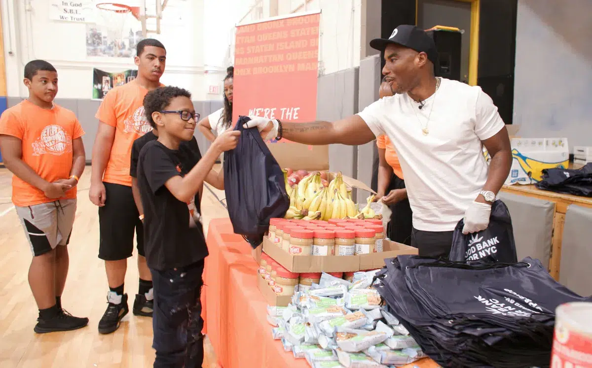 Charlamagne tha God smiles while handing a young boy with glasses a bag filled to the brink with healthy food.