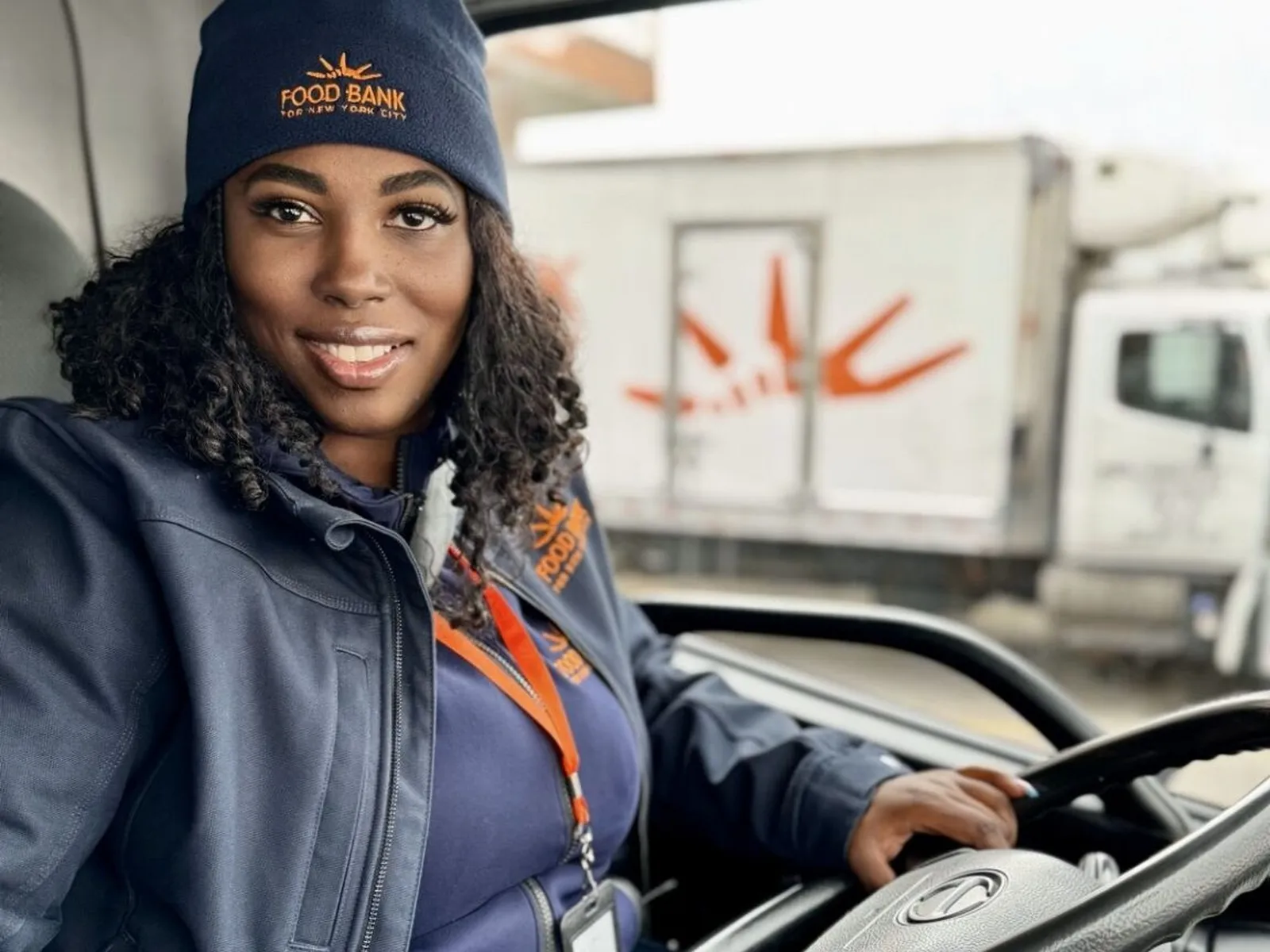 Food Bank driver in delivery truck Smiling Food Bank for NYC driver wearing branded beanie and jacket, sitting in the driver’s seat of a truck with another Food Bank truck in the background.