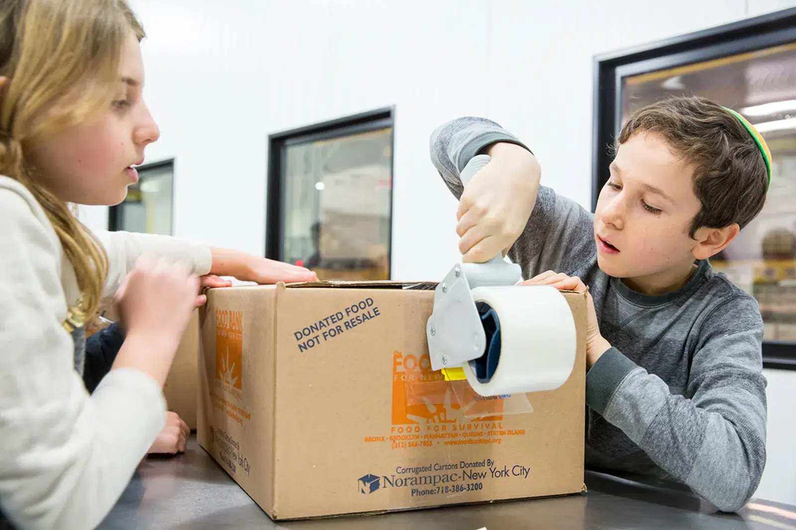 Kids packing food donation box Two children sealing a Food Bank for NYC donation box with packing tape in a warehouse setting