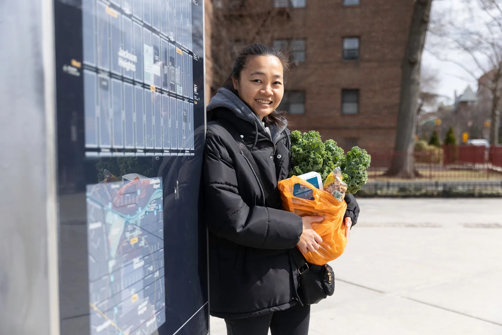 woman with fresh produce” Alt text: Smiling woman in a black coat holding an orange bag of kale and groceries at an outdoor location