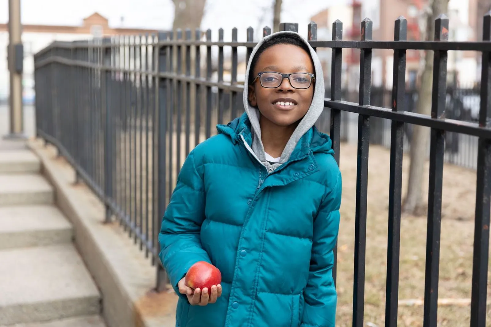 Smiling boy in teal winter coat and glasses holding a red apple, standing next to a black metal fence.
