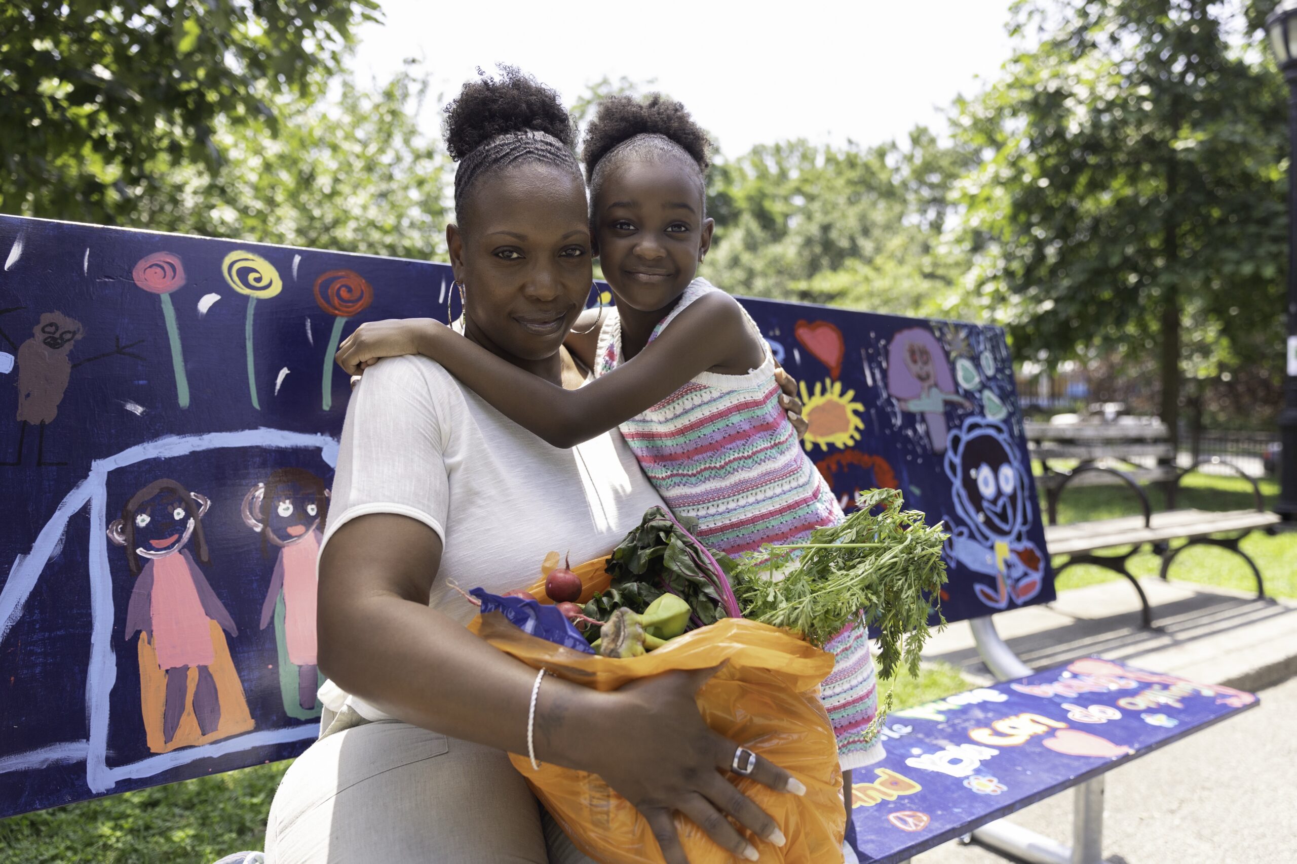 Young girl embraces her mom as they hold bags filled with fresh produce from Food Bank For NYC