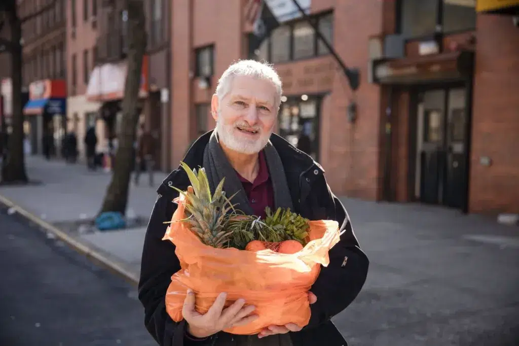 Smiling man standing on a city street holding an orange bag filled with pineapples and other fresh produce.