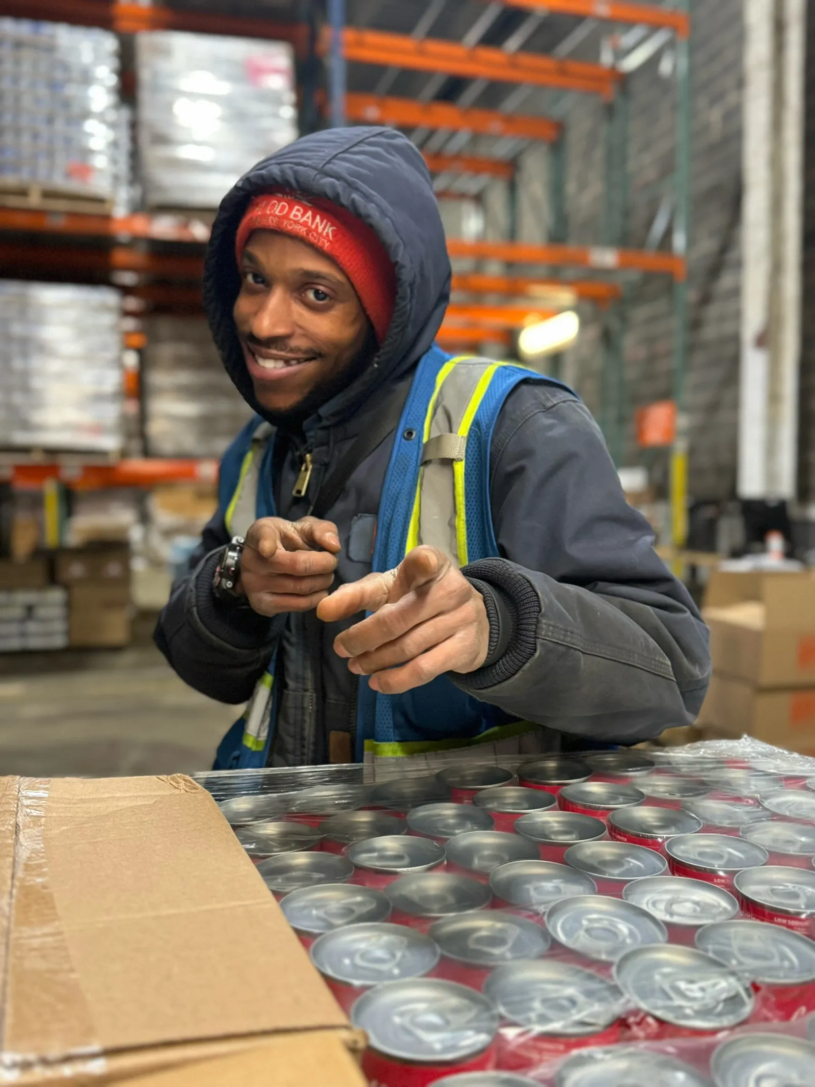 Marcus_ Warehouse worker in Food Bank beanie pointing at camera beside canned goods