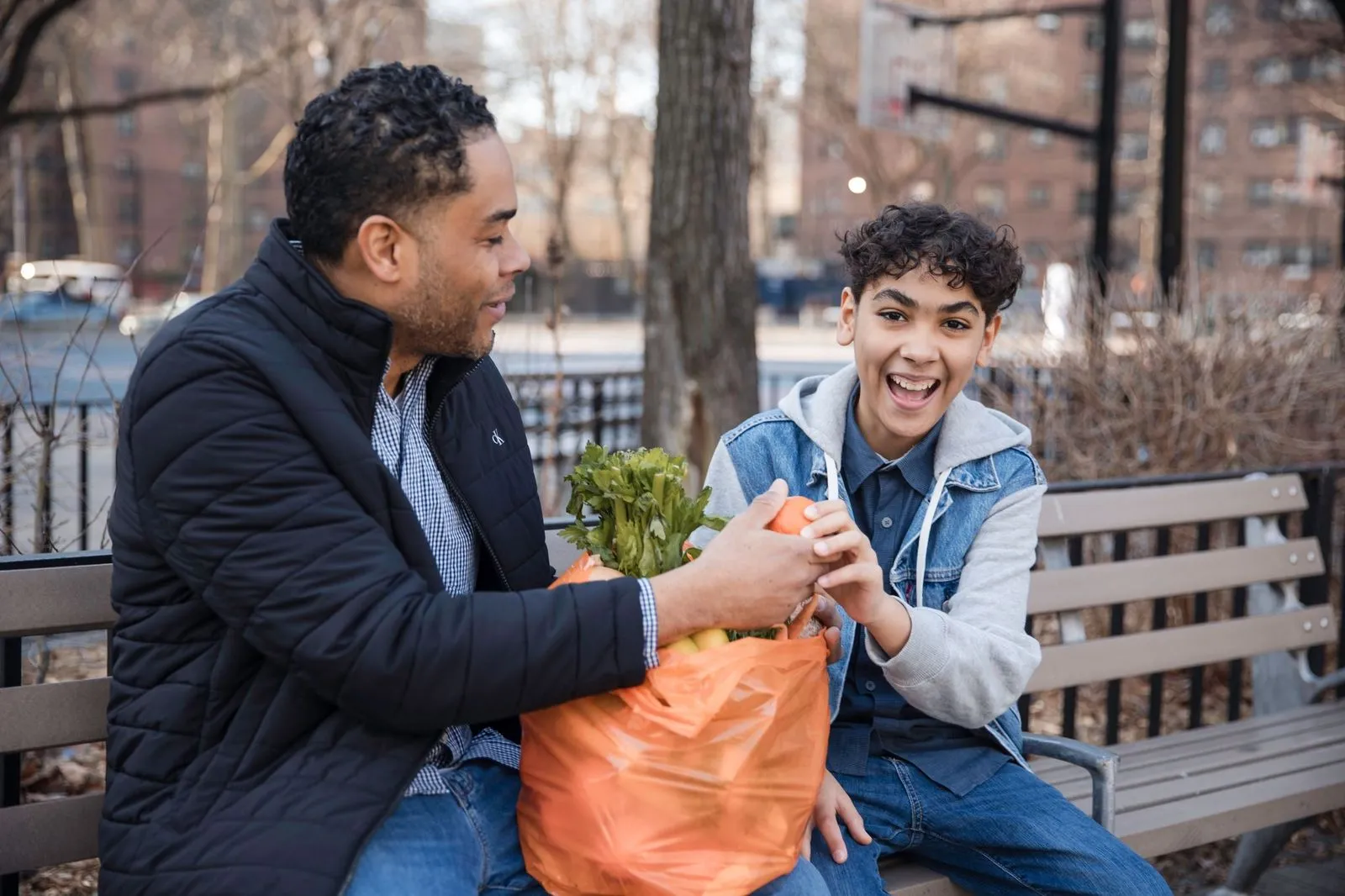 Man and boy smiling on park bench with bag of fresh produce.