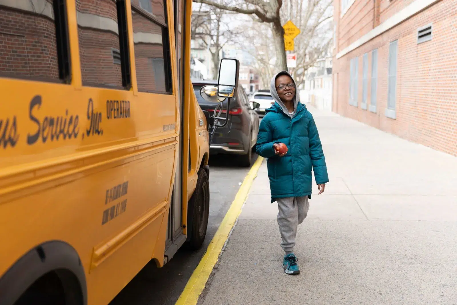 School bus_ Smiling child wearing glasses and a teal winter coat walking on a sidewalk next to a yellow school bus, holding an apple in one hand
