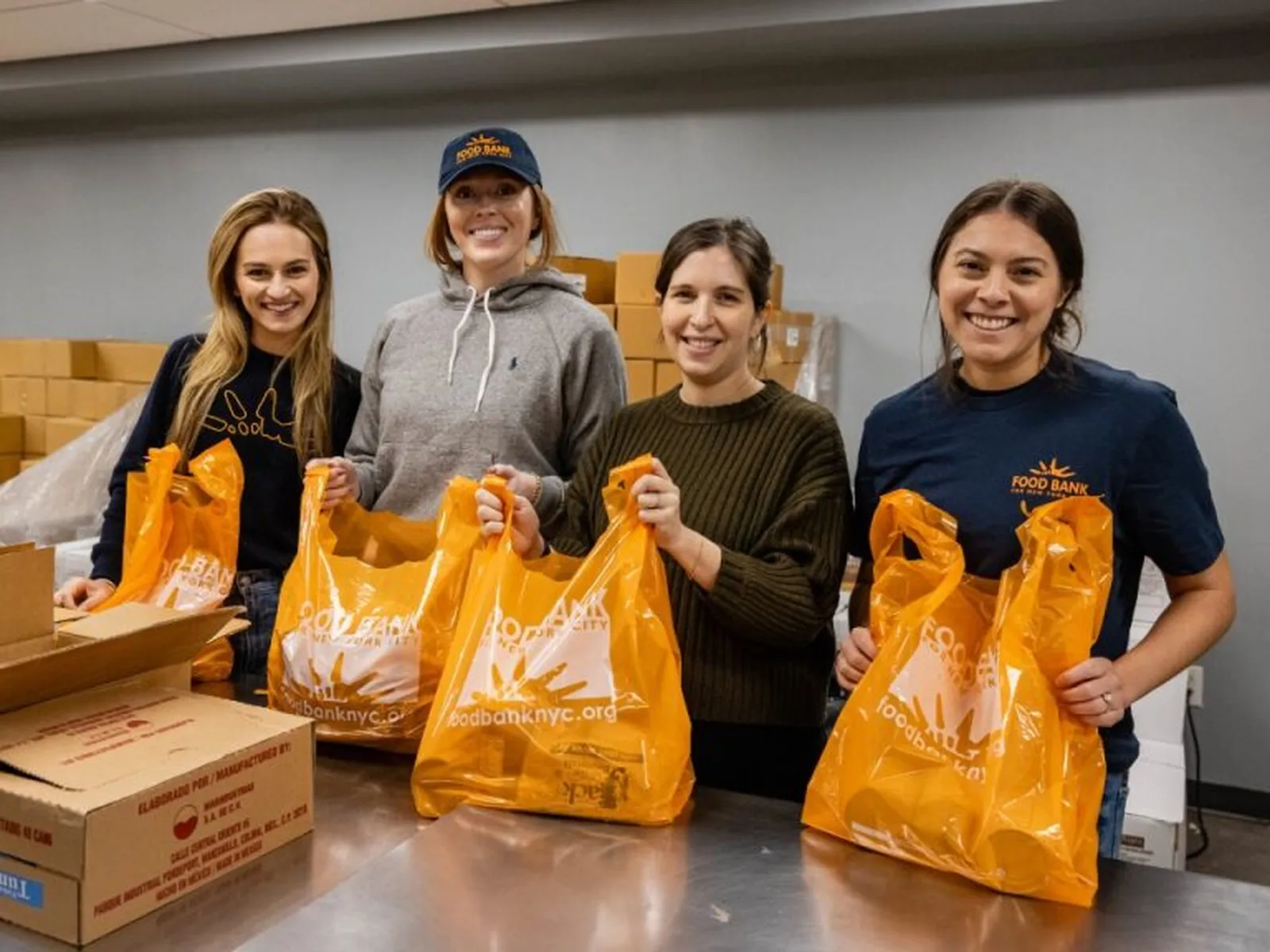 Four smiling volunteers holding orange Food Bank for NYC bags filled with groceries in a warehouse setting.