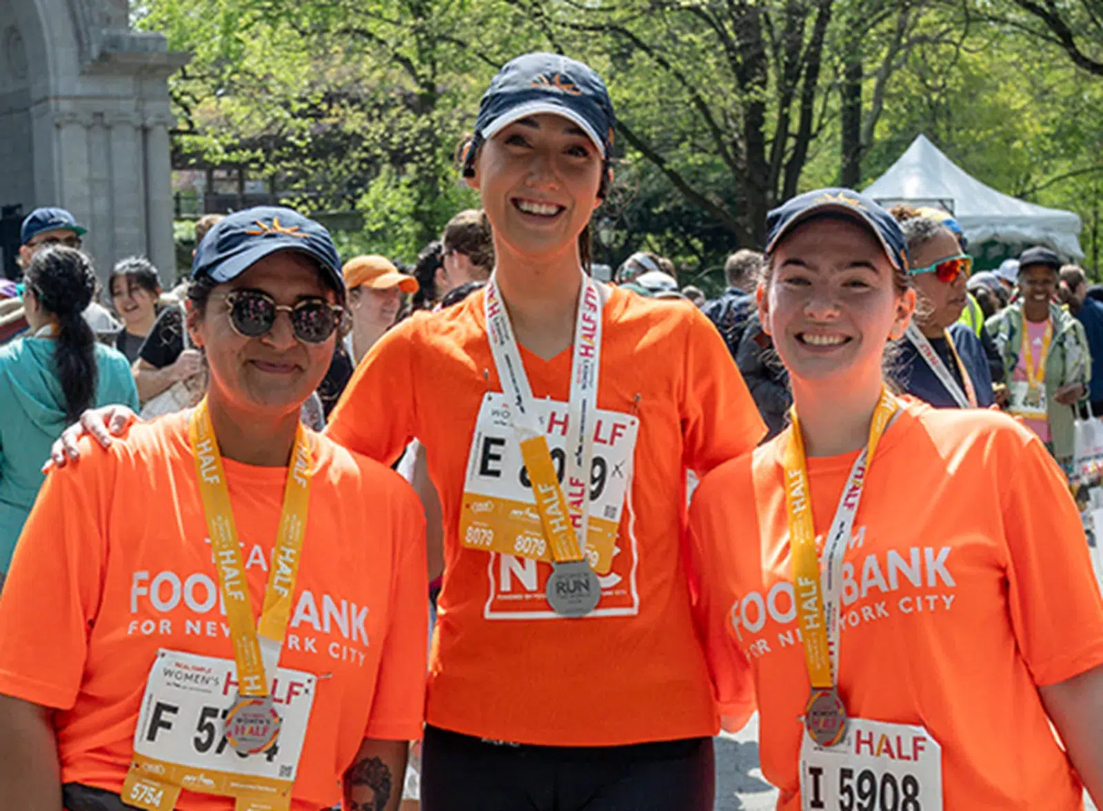 Three smiling runners in orange Food Bank for NYC shirts with medals after a half marathon.