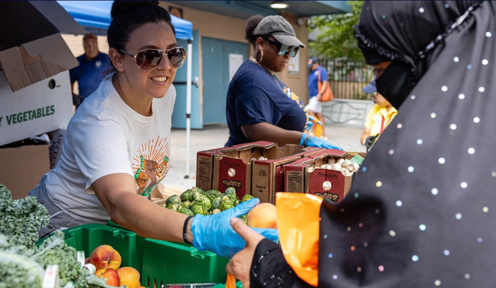 Smiling volunteer in sunglasses and gloves handing a peach to a person at an outdoor produce distribution table