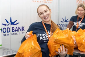 Lauren Patterson, Director, Leadership Giving at Food Bank For NYC smiles at the camera while holding two packed bags of fresh produce for TSA workers.